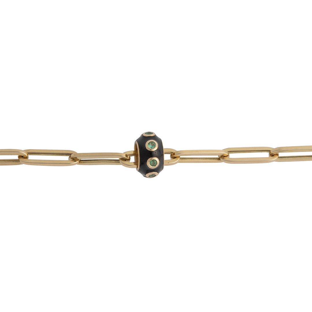A 14k gold paperclip bracelet with a black enamel and emerald bead on a white background