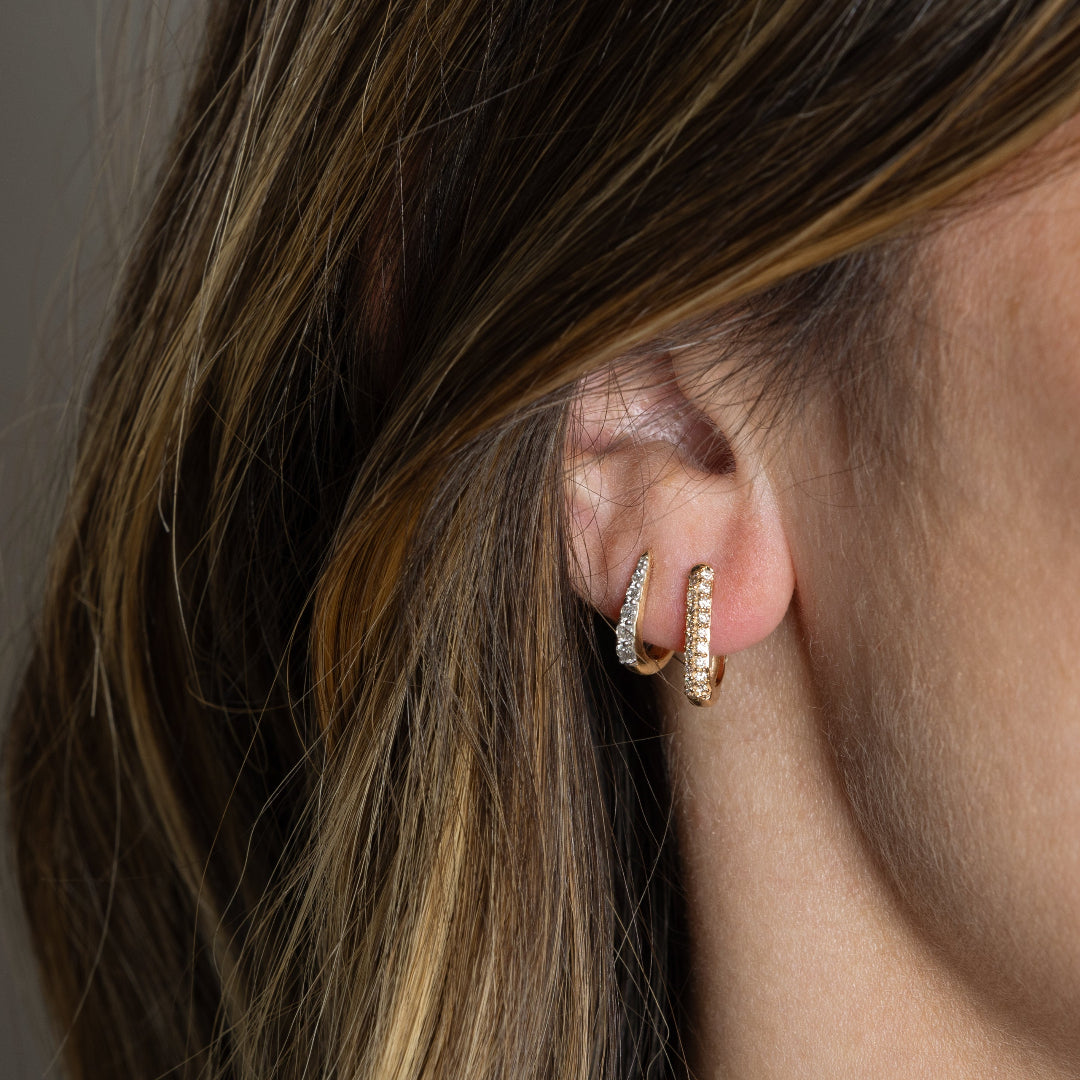 Two 14k gold earrings, one Claw Diamond Huggie Earring with a V-shaped design and diamond embellishments and one traditional diamond pave Huggie Earring on someone's ear.