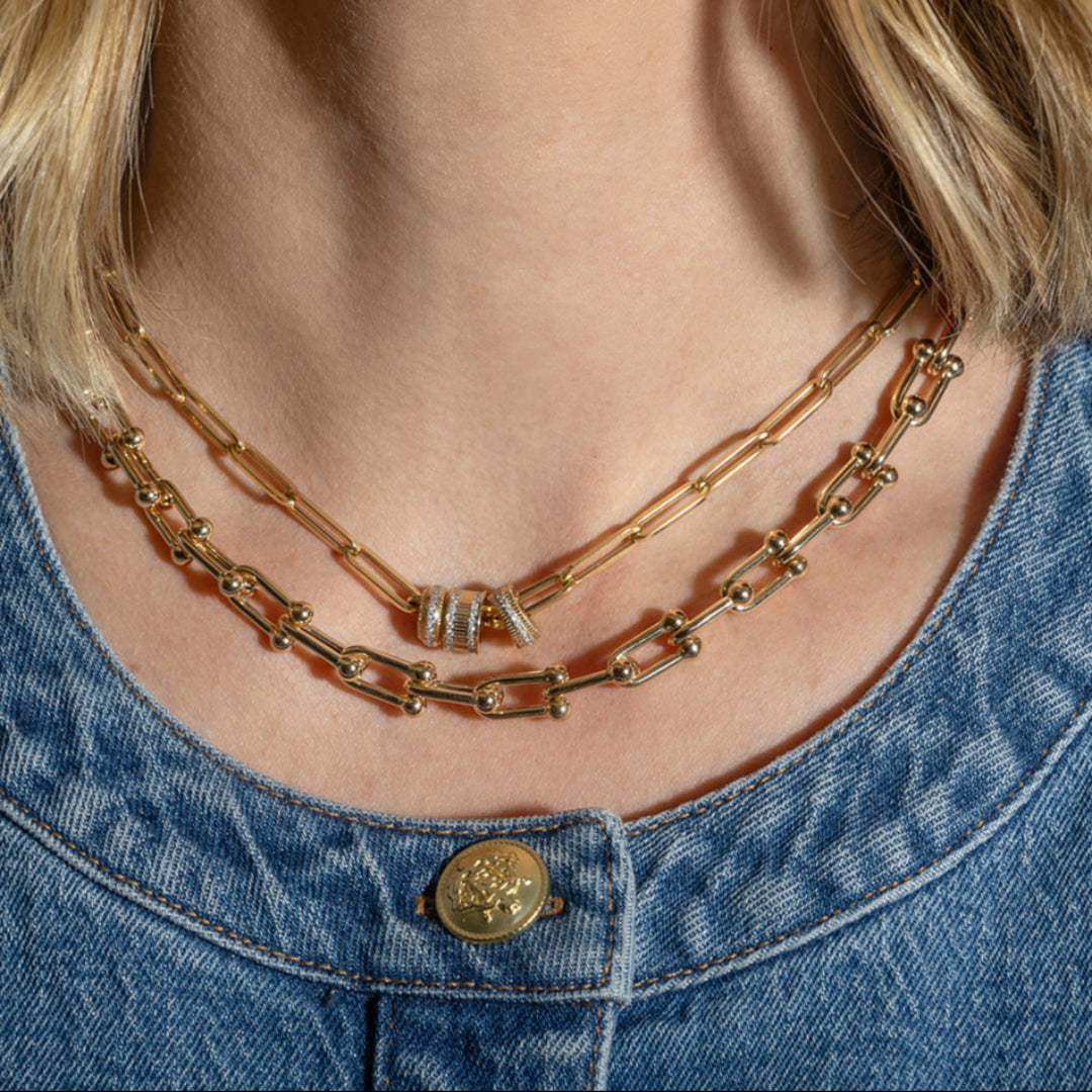 A person wearing two 14k gold necklaces, one is a paperclip chain with three gold and diamond roundel beads and one is a chunky u-link chain.