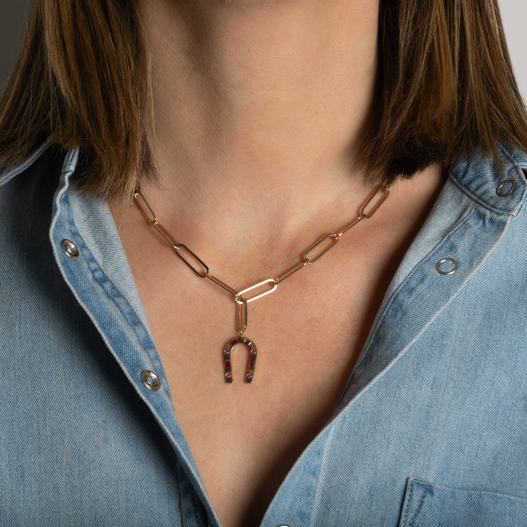 A person wearing a 14k gold paperclip chain with a 14k gold and ruby horseshoe pendant attached to the chain with an elongated oval gold pendant bail.