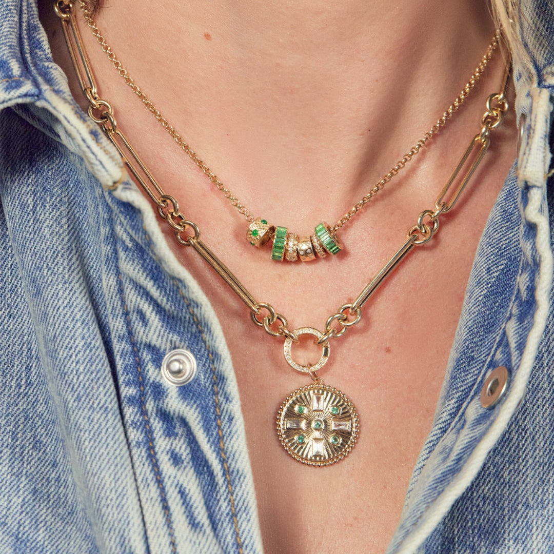 A gold necklace with emerald and diamond beads and a gold link chain with a pendant worn by a person in a denim jacket.