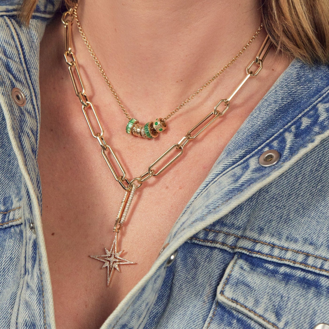 A person wearing two gold necklaces, one with emerald and diamond beads and one with a diamond starburst pendant.