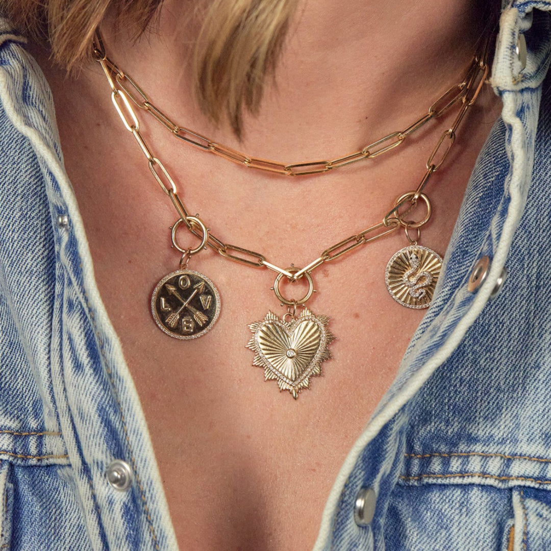 Two gold paperclip chains on a person, with the longer gold paperclip chain featuring three differently styled pendants attached with round dotted pendant bails.