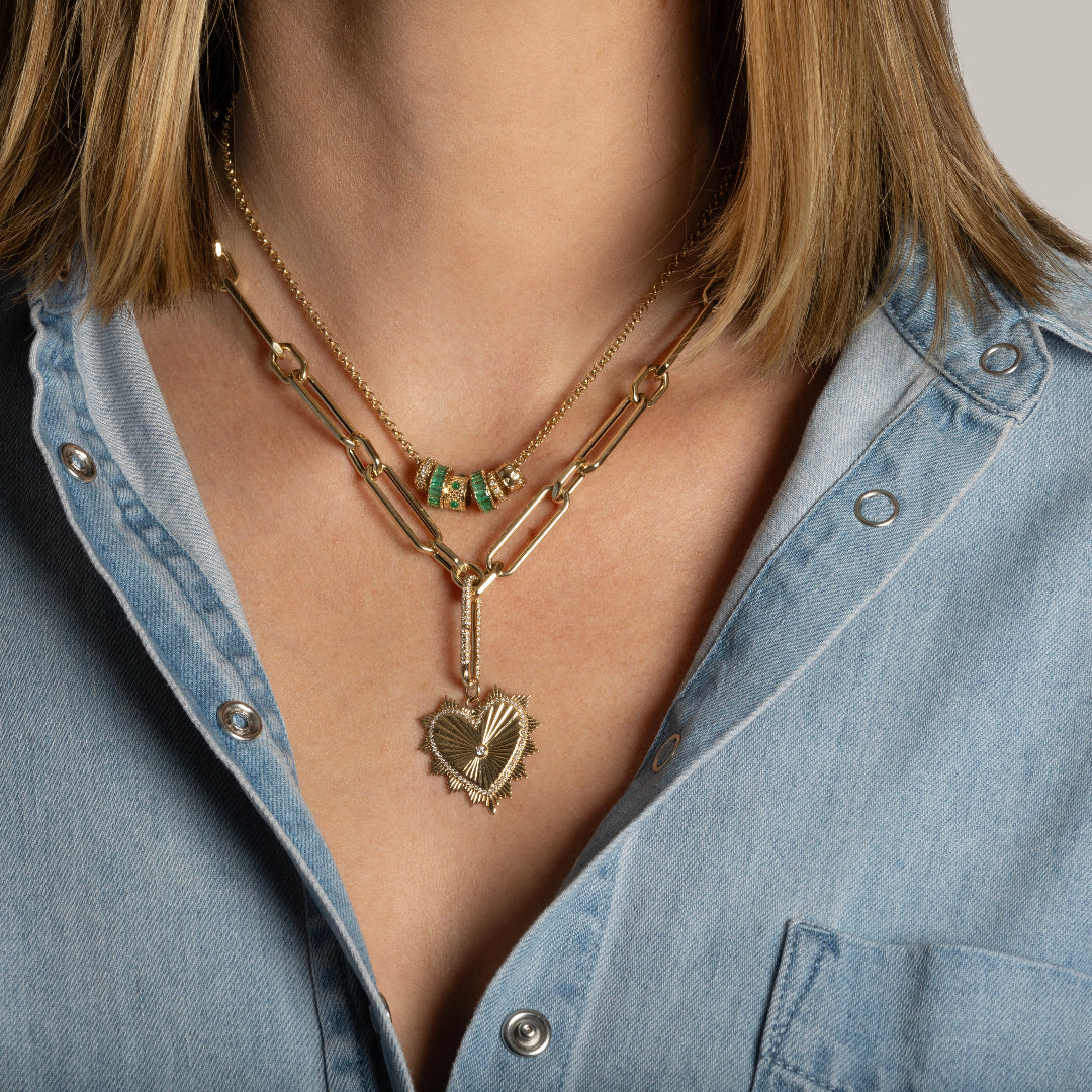 A person wearing a denim shirt with 14k gold hexagon link chunky chain necklace with a heart charm and a diamond link design and a mini rolo chain with diamond and emerald roundel beads.