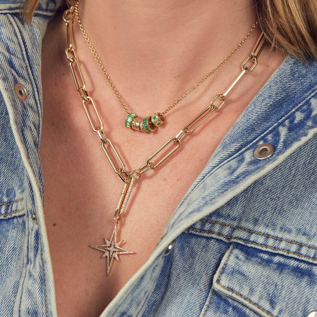 A person wearing a 14k gold hexagon link chunky chain necklace with a diamond link design and a mini rolo chain with diamond and emerald roundel beads.