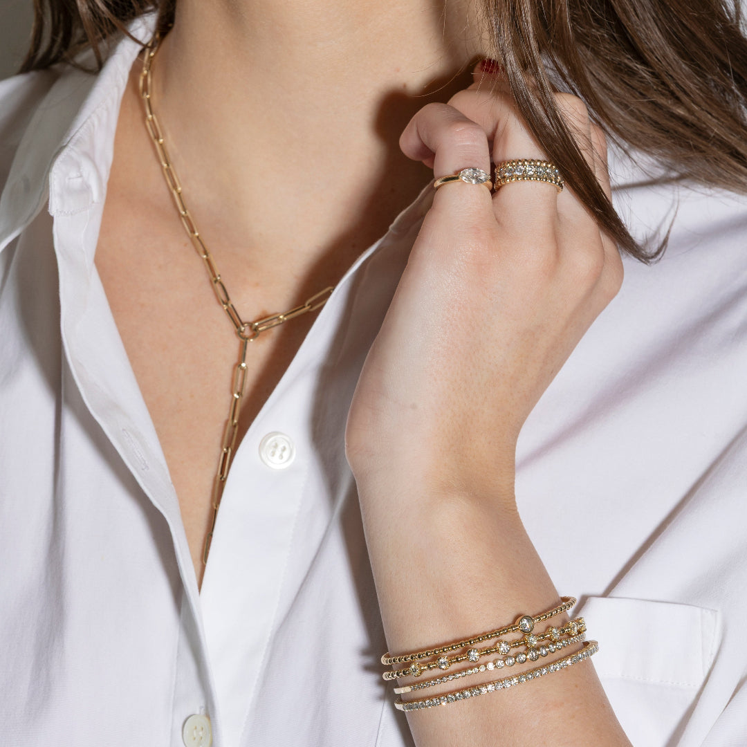Close-up of a person wearing gold jewelry including a necklace, bracelet, and ring on a white shirt background.