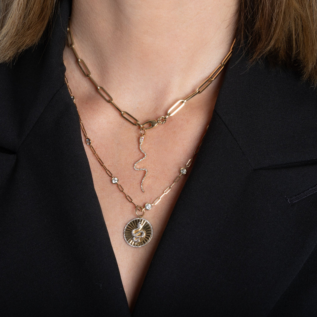 A person wearing a 14k gold paperclip chain necklace with a 14k gold diamond serpent charm layered with a 14k gold diamond station necklace and a 14k gold serpent charm.