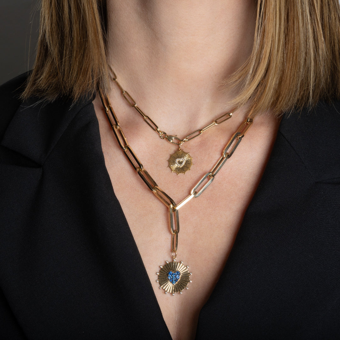 A person wearing two paperclip chain necklaces; one chain has a gold and diamond starburst pendant and the other chain is chunkier and styled lariat-style with a circle pendant featuring a blue sapphire heart.