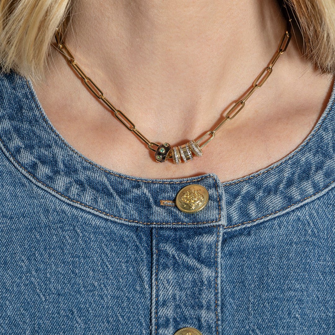 A person wearing a 14k gold paperclip chain necklace with three diamond and gold roundel beads and a black enamel and emerald roundel bead.