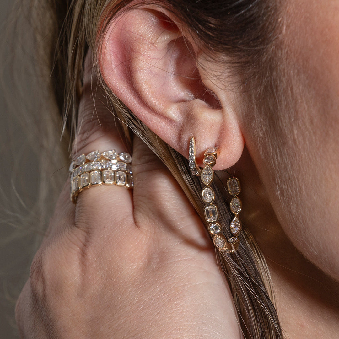 A person pulling their hair back from their face showcasing stacking diamond band rings of different shaped diamonds set in 14k gold and two earrings.