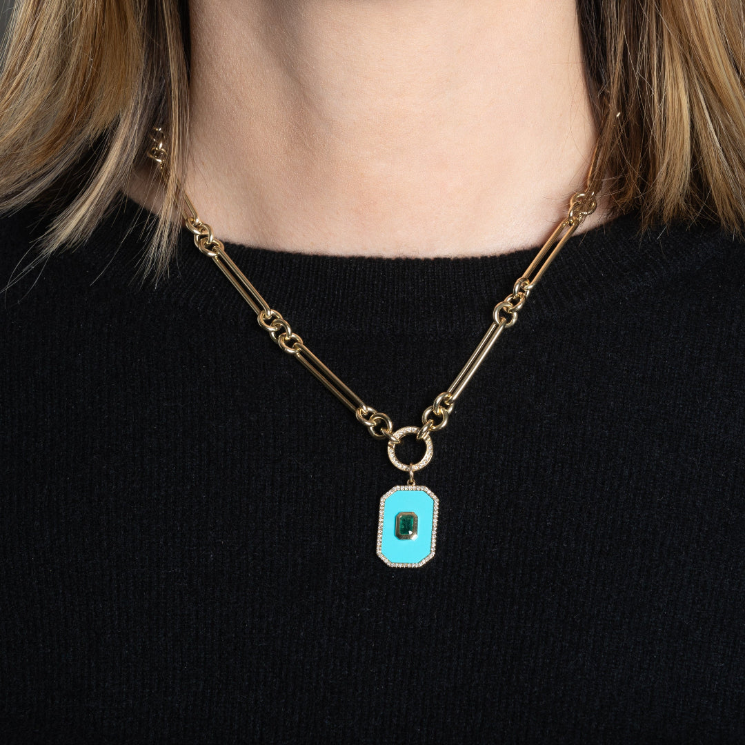 A person wearing a gold chain with a rectangular turquoise, emerald and diamond pendant attached.