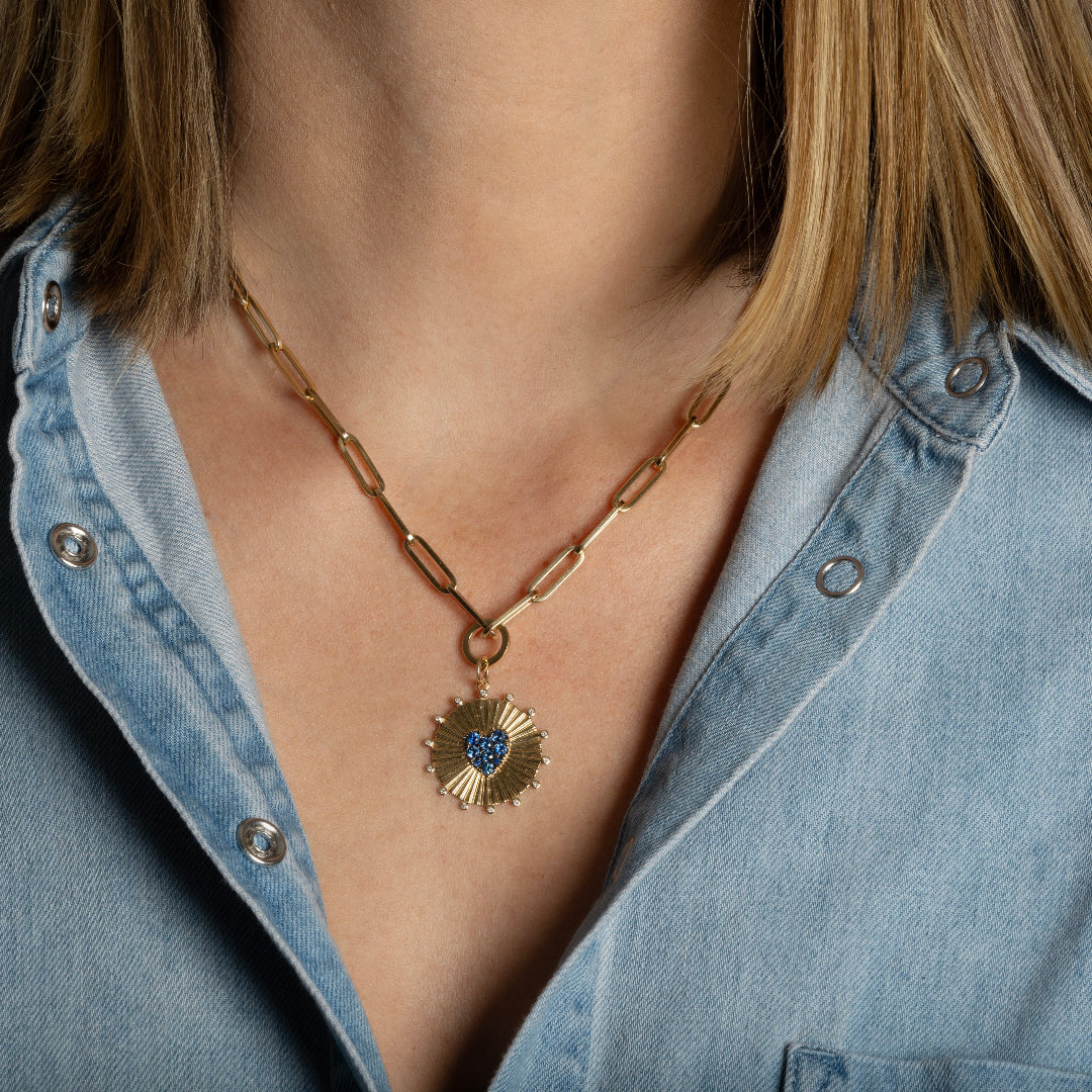 A person wearing a 14k gold paperclip chain necklace with a round pendant bail attaching a round pendant with a blue sapphire heart.