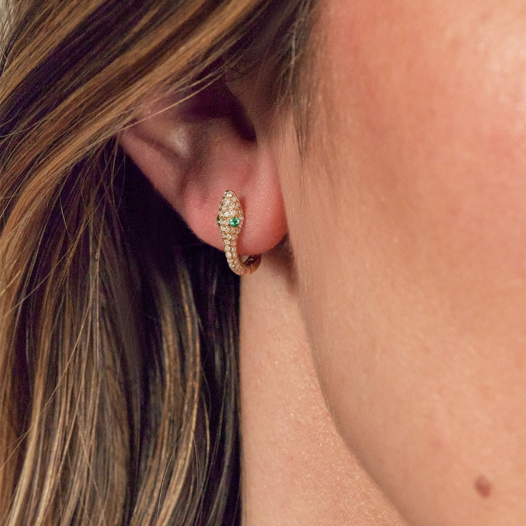 A person wearing gold serpent-shaped huggie earrings adorned with natural diamonds and emeralds.