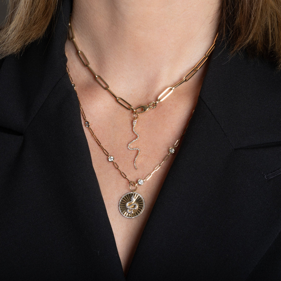 A person wearing a 14k gold paperclip chain necklace with a 14k gold diamond serpent charm layered with a 14k gold diamond station necklace and a 14k gold serpent charm.