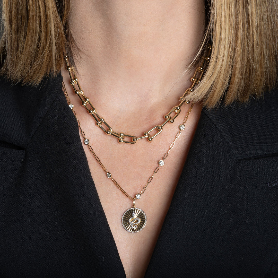 A person wearing a 14k gold paperclip chain necklace with a 14k gold diamond serpent charm layered with a 14k gold diamond station necklace and a 14k gold serpent charm.