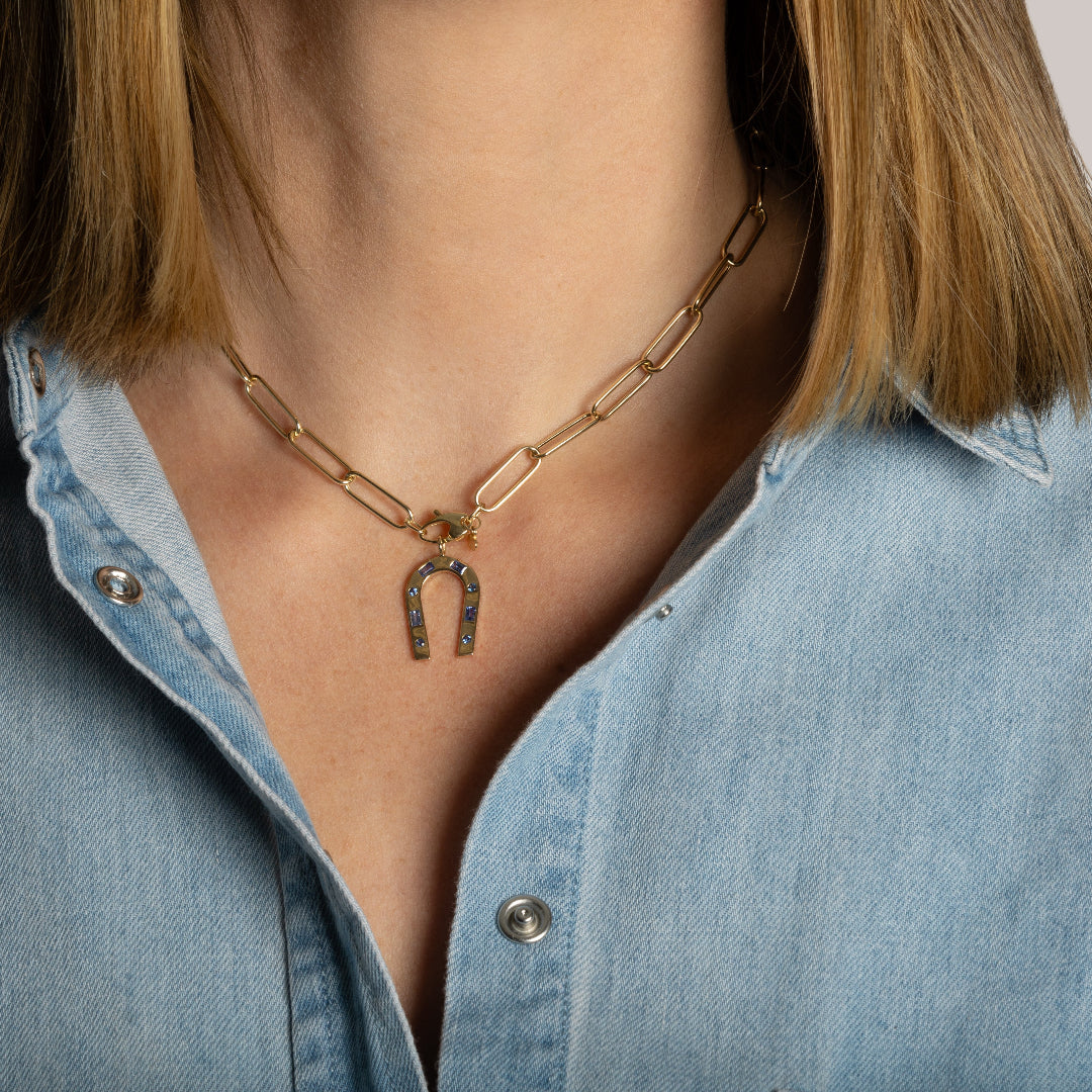 A person wearing a blue denim shirt modeling a 14k gold paperclip chain and a blue sapphire horseshoe charm.