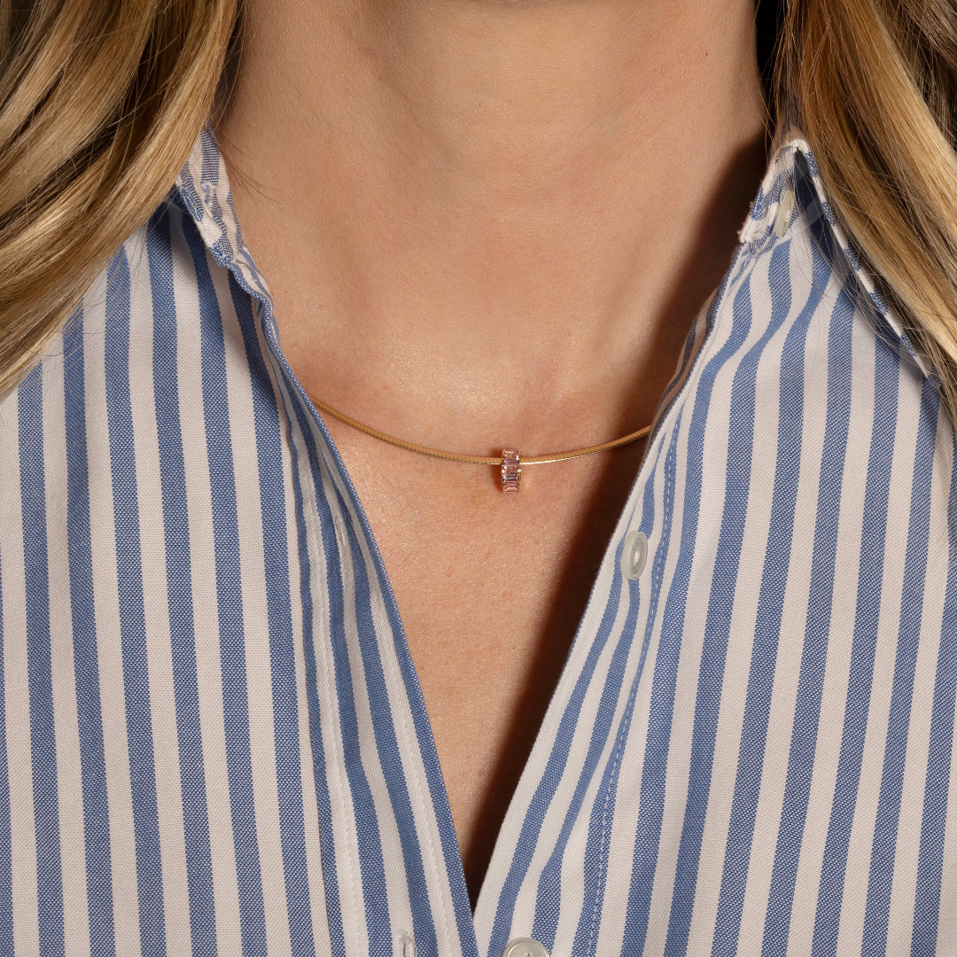 A gold wire collar necklace with a pink bead worn by a person in a blue and white striped shirt.