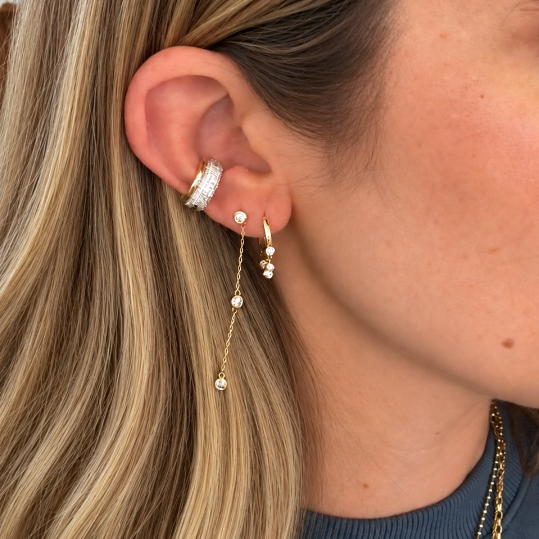 A close-up of a woman's ear wearing a gold huggie hoop earring, a small gold stud with a dangling chain and two ear cuffs. She also has layered gold necklaces, against her blonde hair and a blue-gray top.