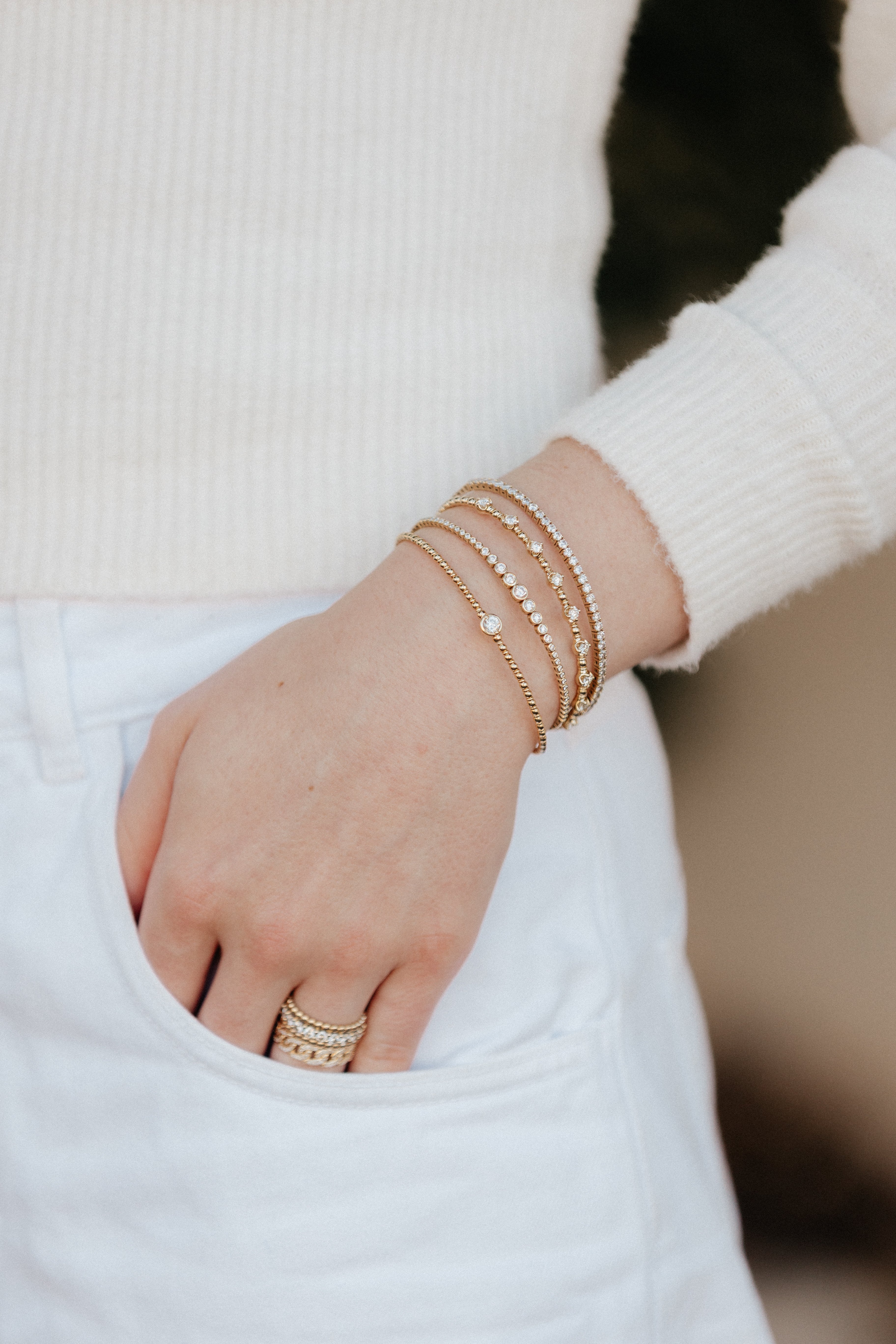 A person wearing a cream sweater and white jeans, showcasing a wrist full of gold bracelets. 