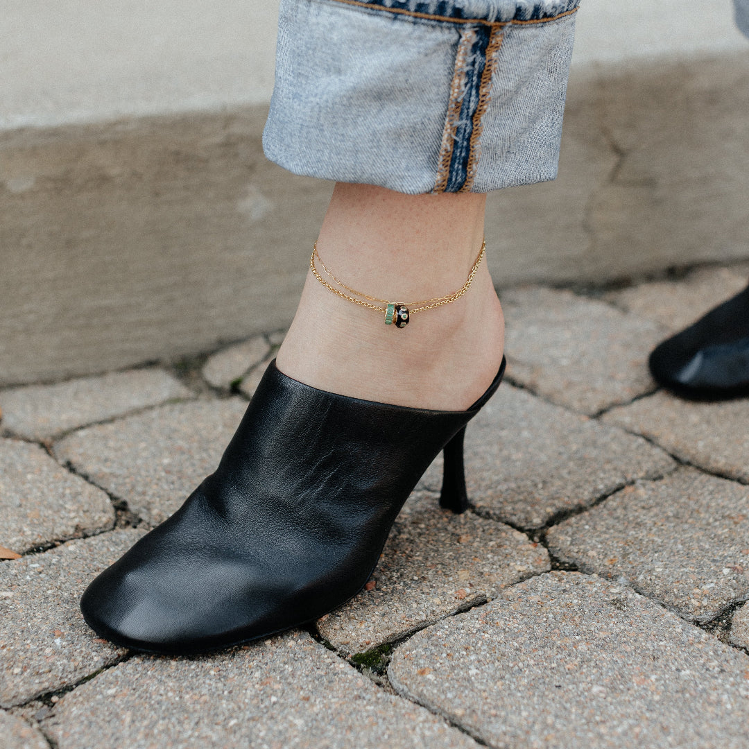 Two ankle bracelets worn with rolled-up jeans on a stone pavement.