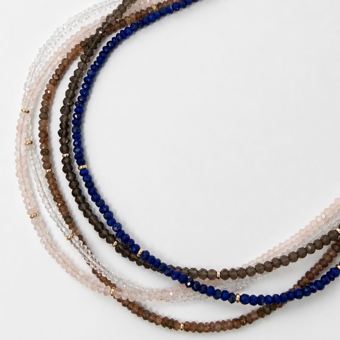 A set of multi-strand beaded necklaces arranged in a circle on a plain white background. The necklaces feature faceted beads made from lapis, smoky quartz, andalusite, white topaz and rose quartz separated by small 14k gold spacer beads.