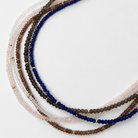 A set of multi-strand beaded necklaces arranged in a circle on a plain white background. The necklaces feature faceted beads made from lapis, smoky quartz, andalusite, white topaz and rose quartz separated by small 14k gold spacer beads.