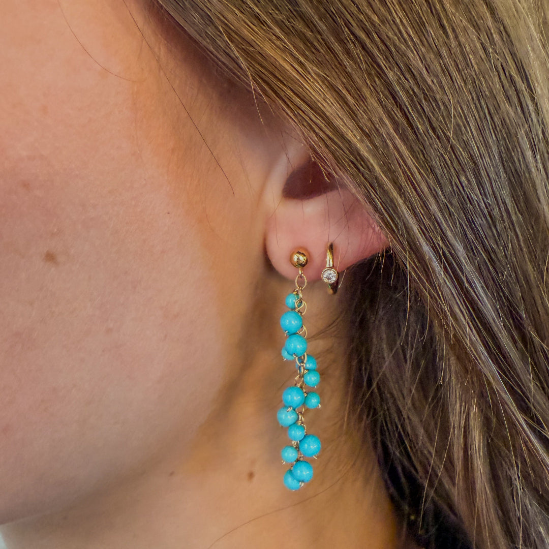 Close-up of a person wearing turquoise drop earrings and gold bezel set diamond huggie earrings. 