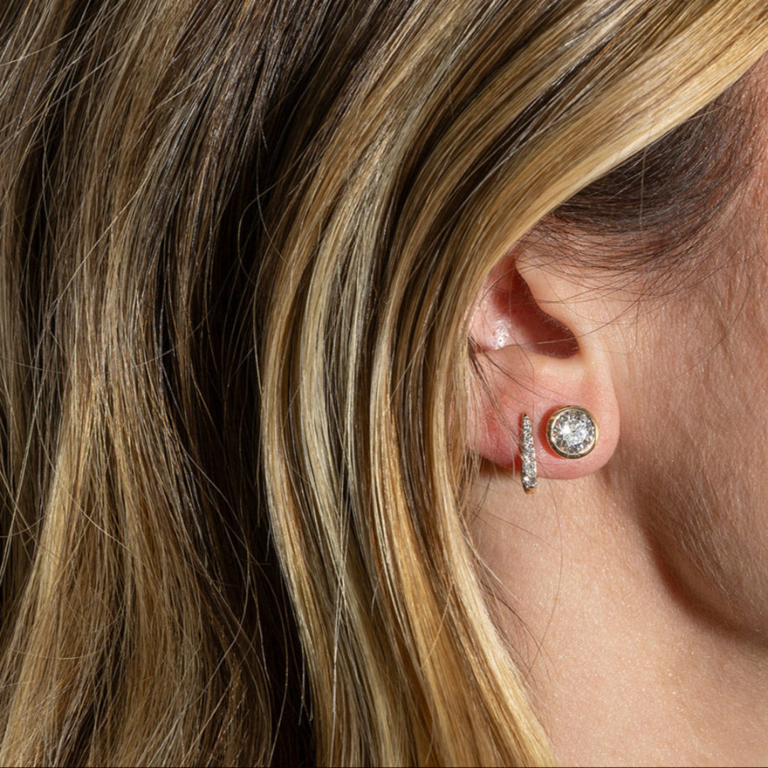 A close-up image of a woman's ear wearing a round diamond stud earring.