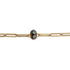 A 14k gold paperclip bracelet with a black enamel and emerald bead on a white background