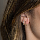 Two 14k gold earrings, one Claw Diamond Huggie Earring with a V-shaped design and diamond embellishments and one traditional diamond pave Huggie Earring on someone's ear. 