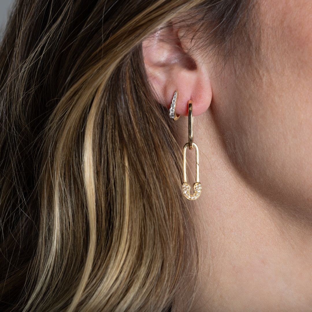 Close-up of an ear wearing gold hoop earrings, one claw shaped huggie and one lager hoop with a diamond studded safety pin. 