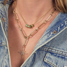 A person wearing two gold necklaces, one with emerald and diamond beads and one with a diamond starburst pendant.