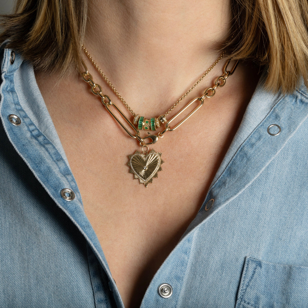 A person wearing a mini rolo chain with diamond and emerald roundel beads and a  chunky 14k gold chain with a beveled heart pendant. 