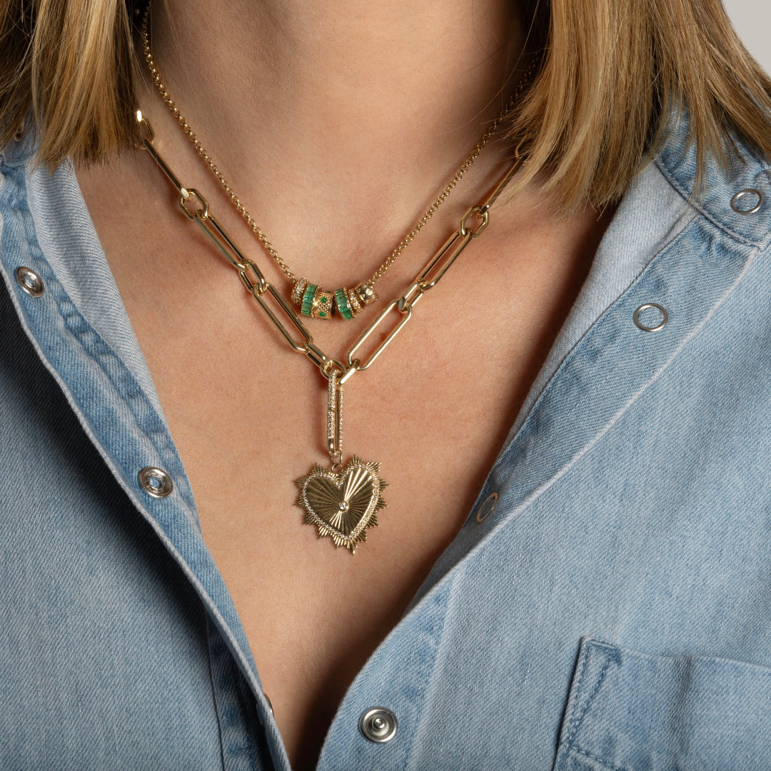 A person wearing a mini rolo chain with diamond and emerald roundel beads and a  chunky 14k gold lariat chain with a beveled heart pendant attached. 