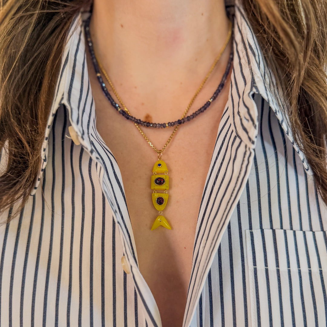 Close-up of a person wearing a striped shirt with layered necklaces, including an iolite beaded necklace and a 14k gold chain with a yellow quartz fish-shaped pendant.