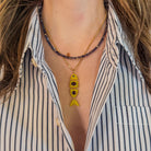 Close-up of a person wearing a striped shirt with layered necklaces, including an iolite beaded necklace and a 14k gold chain with a yellow quartz fish-shaped pendant.