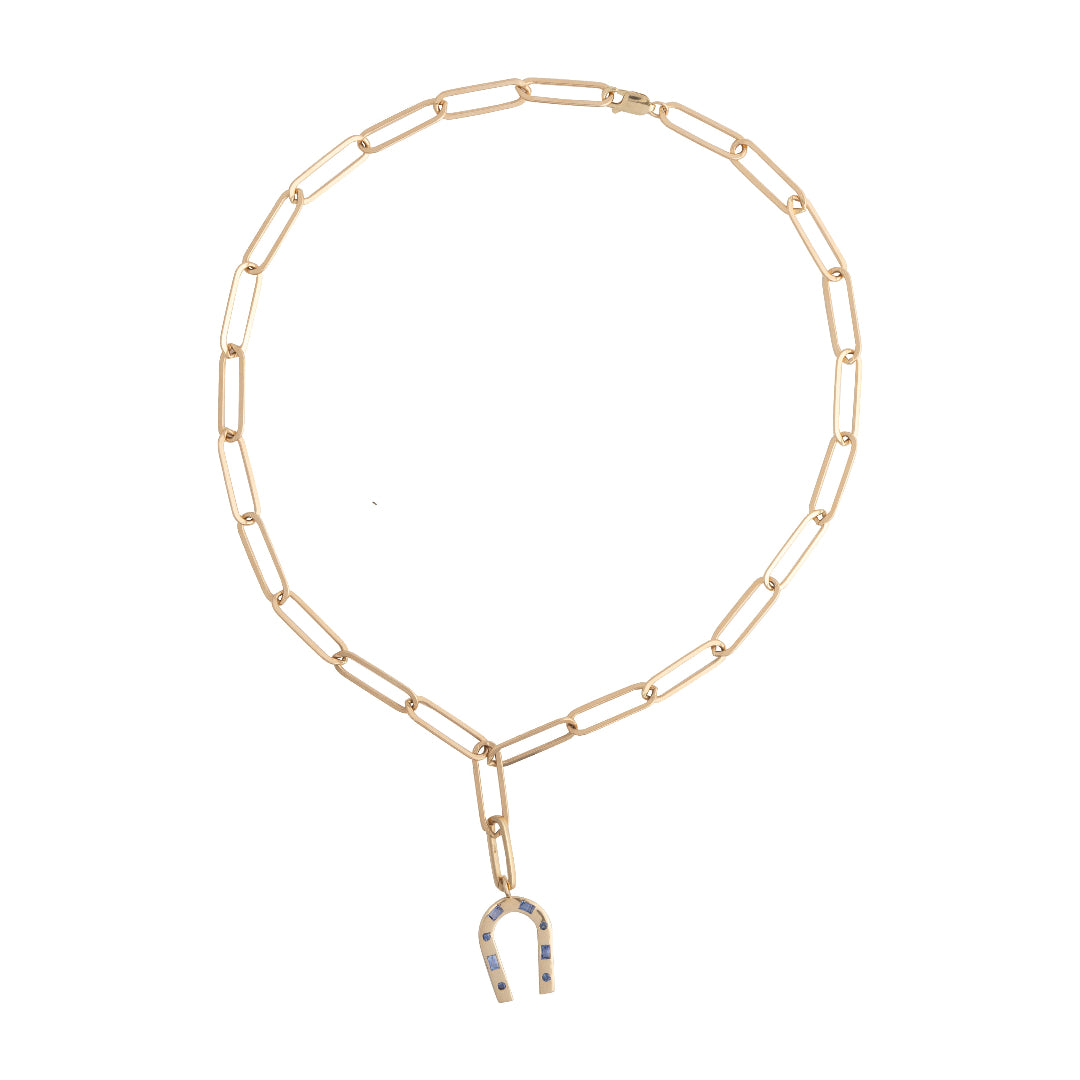 A 14k gold paperclip chain styled lariat style with a blue sapphire encrusted horseshoe charm laid out on a white background. 