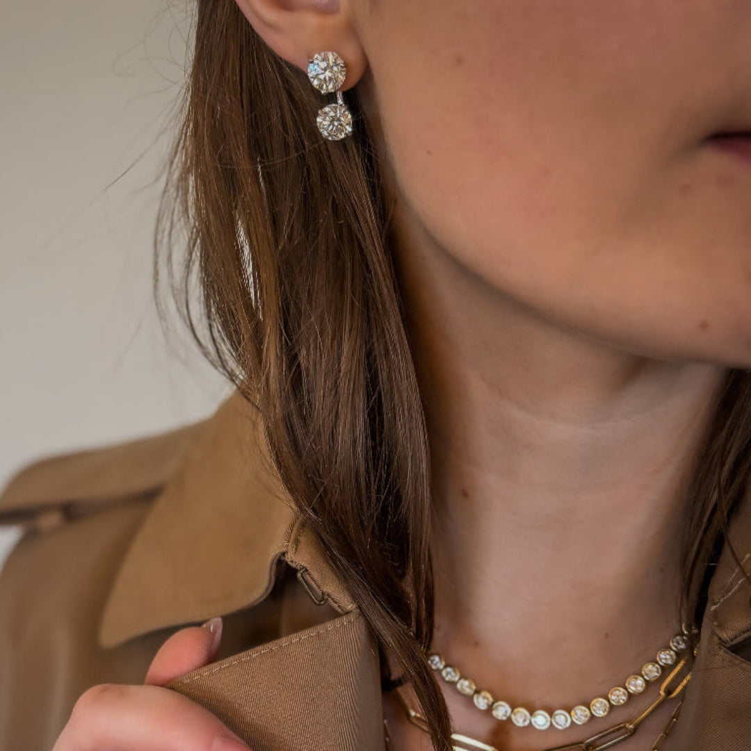 Close-up of a person wearing a beige jacket, gold necklace, and earrings with a neutral background.