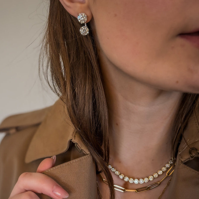 Close-up of a person wearing a beige jacket, gold necklace, and earrings with a neutral background.