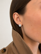A close-up of a model's ear wearing a lab-grown stud in a 14k white gold martini setting, paired with a brown coat against a light neutral background.