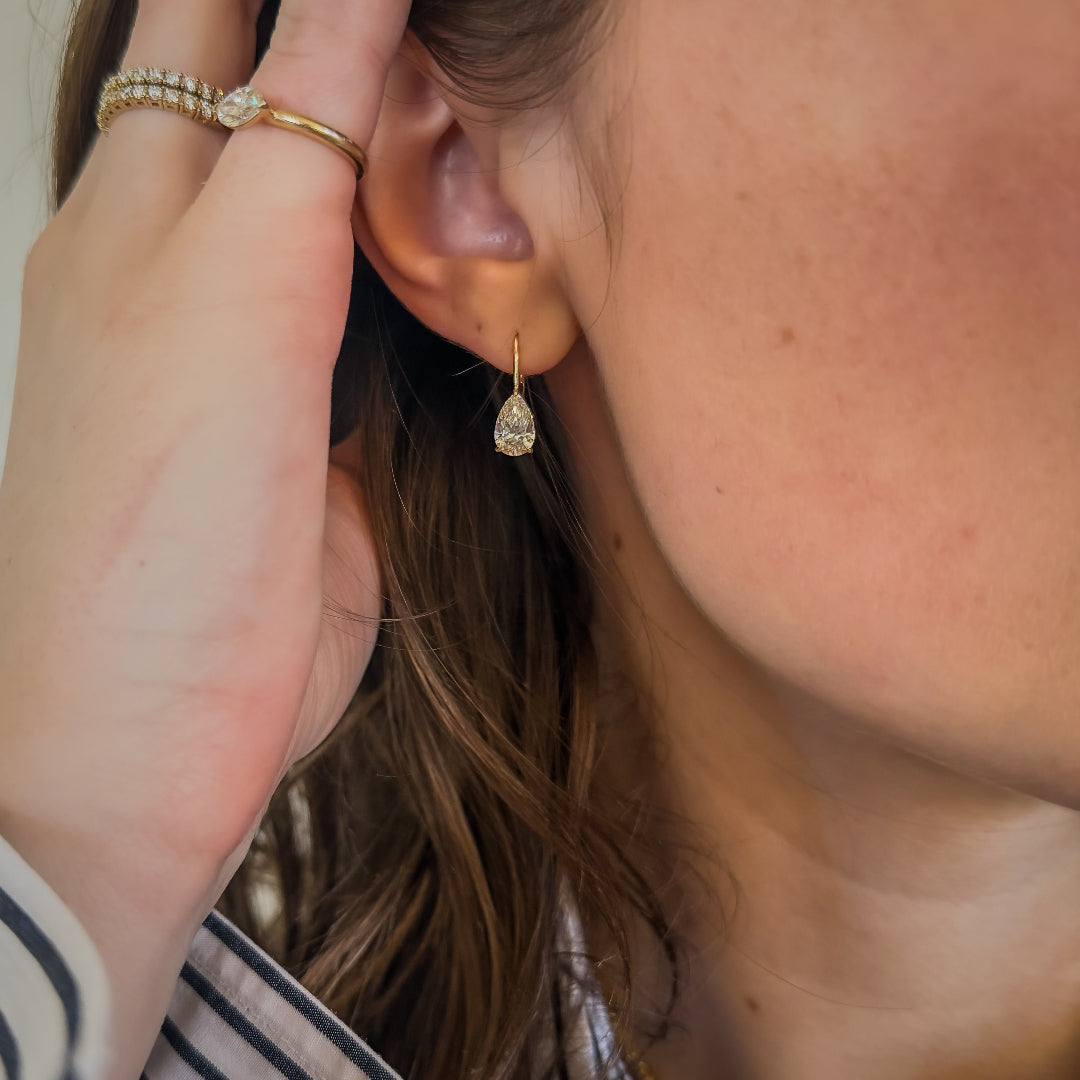 A close-up of a person wearing pear shaped diamond drop earrings set in 14k gold, with their hand near their ear. They are also wearing gold rings.
