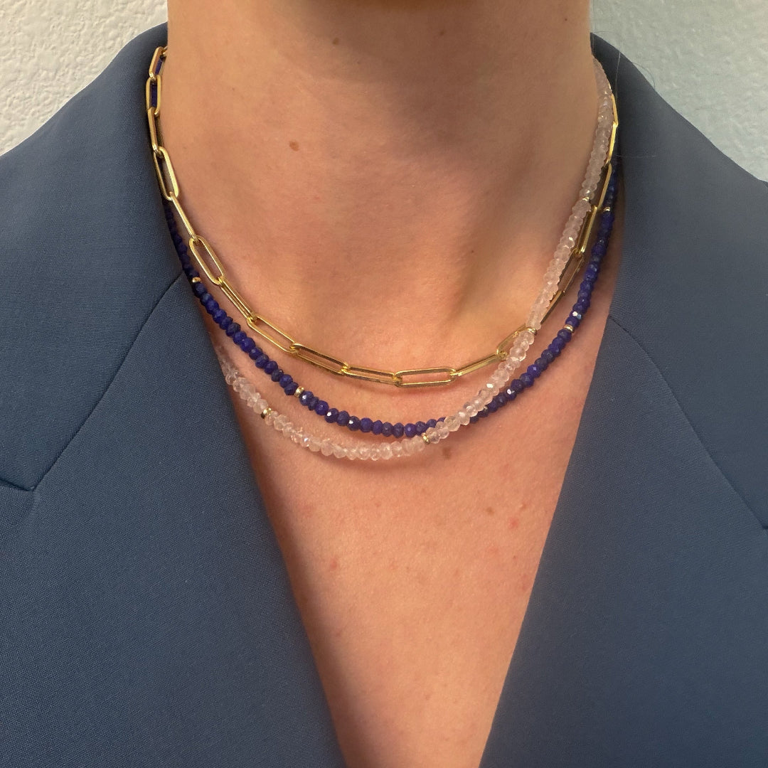 A person wearing two gemstone necklaces made of lapis and rose quarts and a 14k gold paperclip chain. She is also wearing a blue blazer. 
