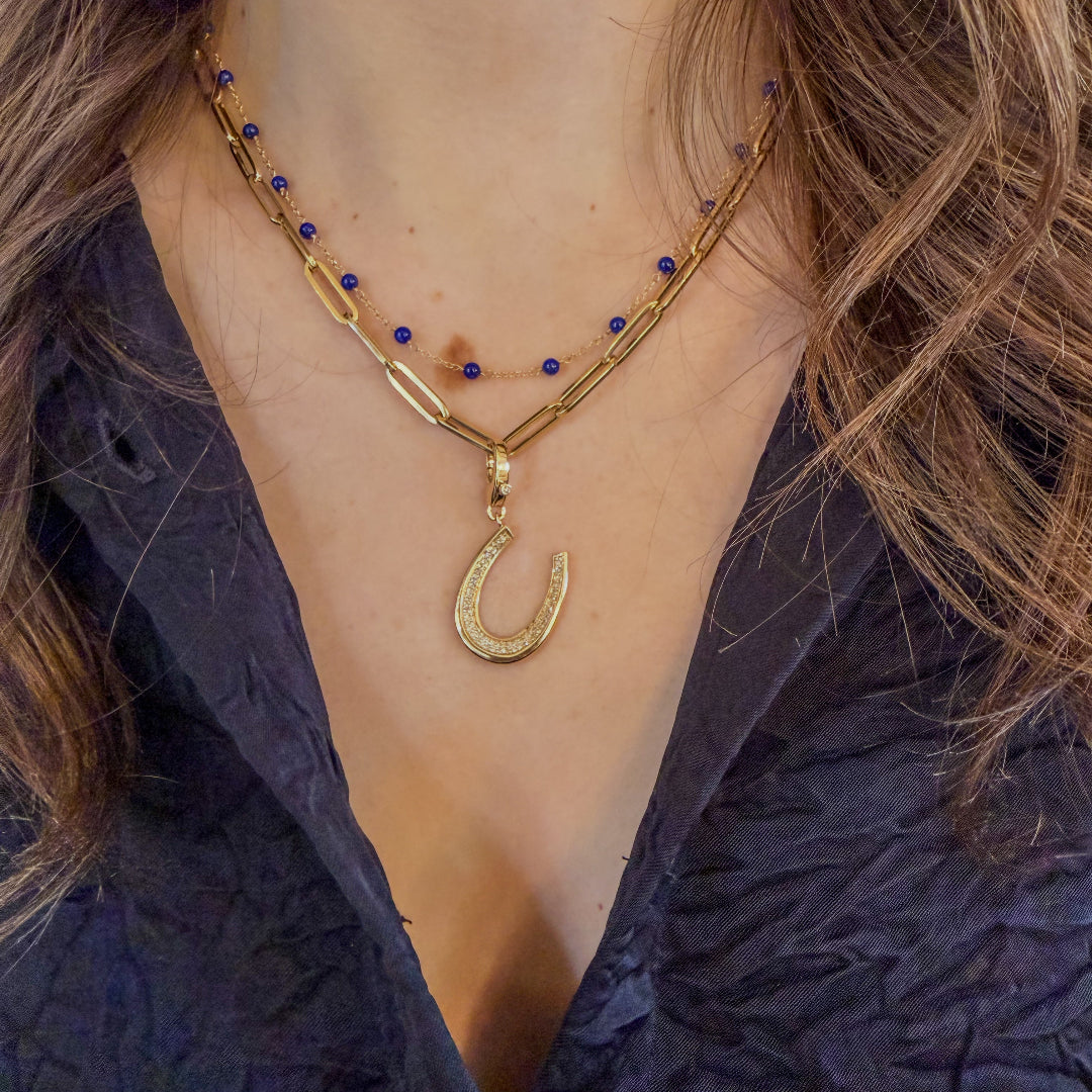 A person with a blue shirt wearing a 14k gold station necklace with lapis heads and a 14k gold paperclip chain with a diamond horseshoe pendant. 