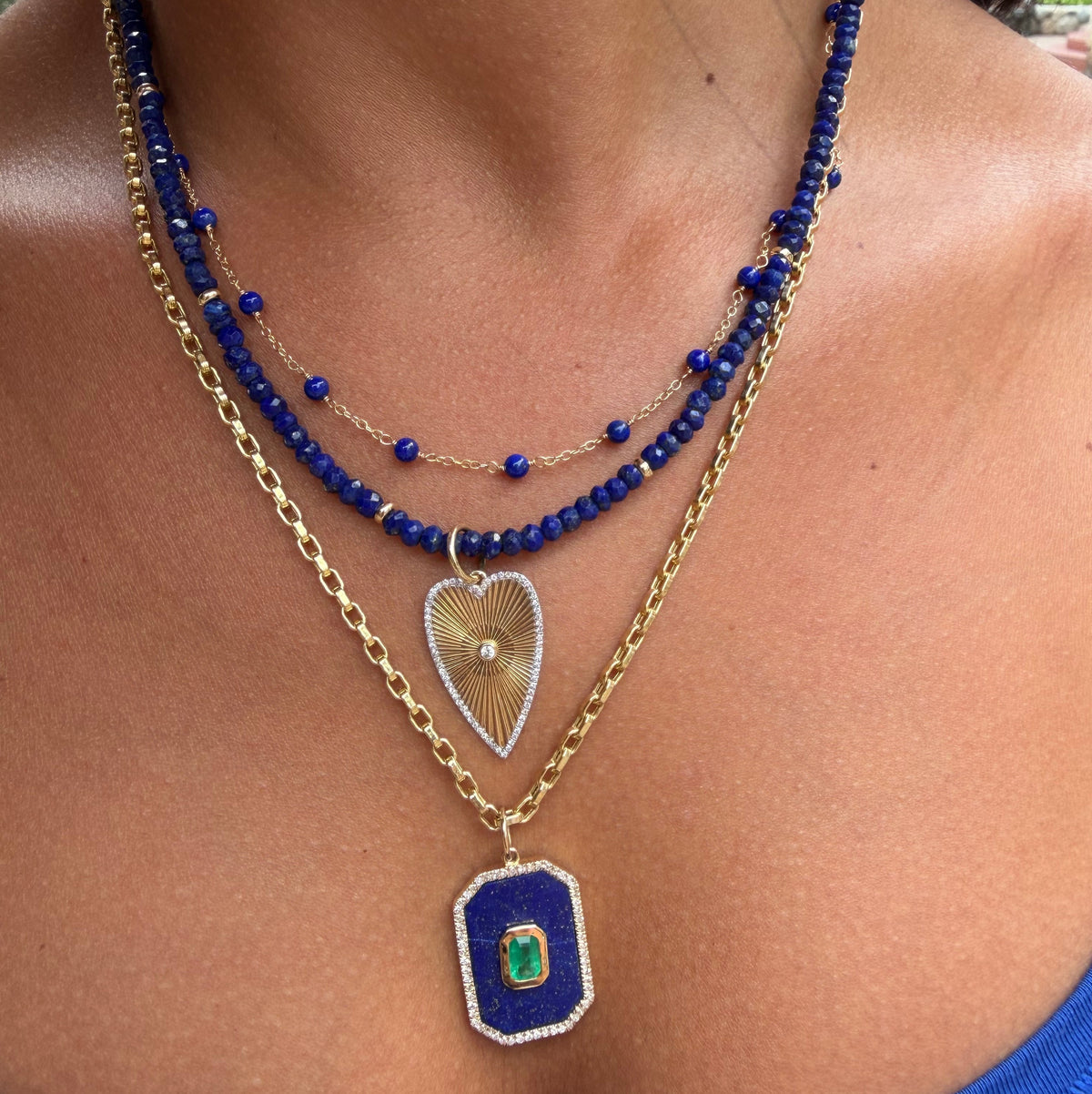 A close-up of a person's chest wearing a layered necklace with a gold chain, blue lapisbeaded strands, a gold teardrop pendant, and a lapis rectangular pendant with an emerald.