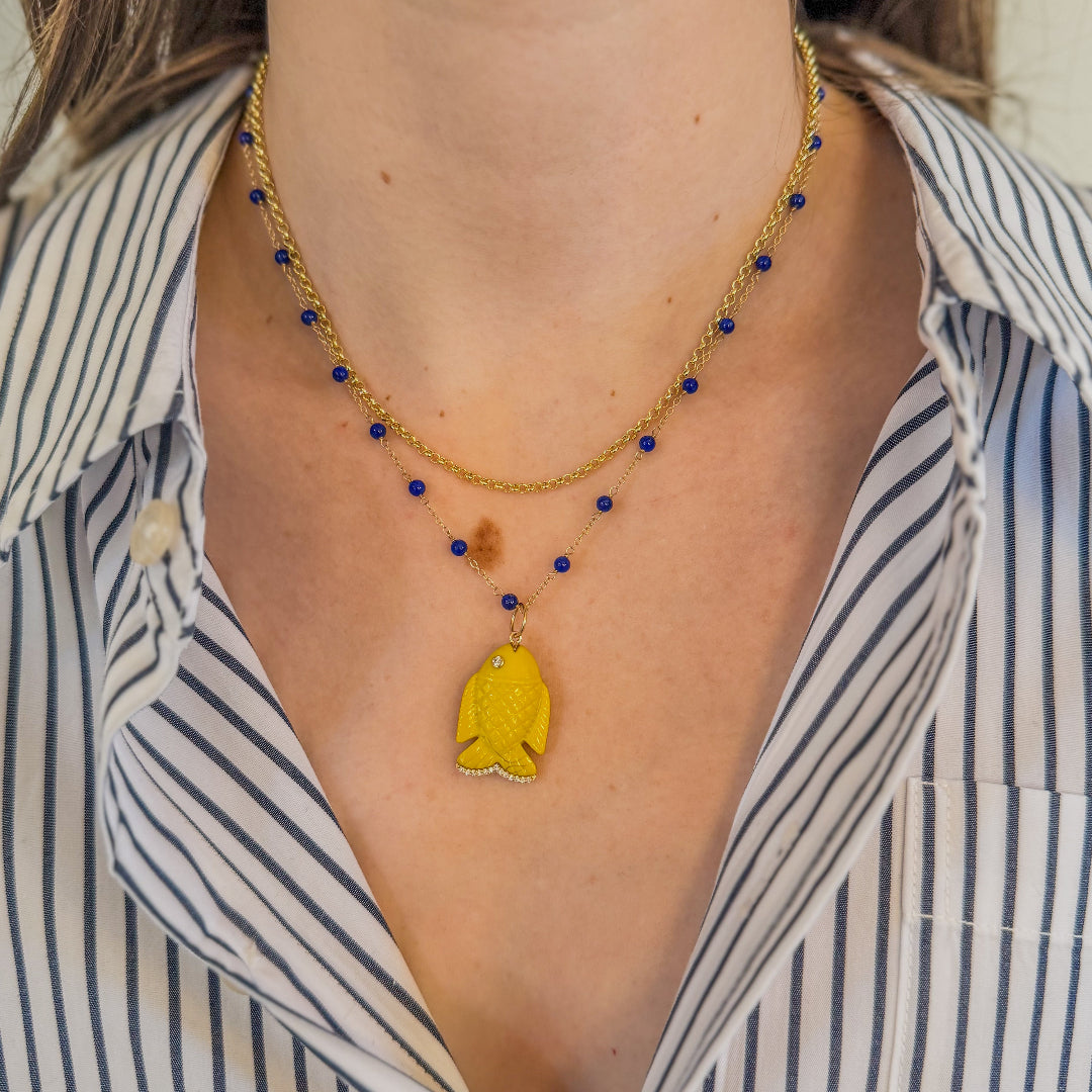 Person wearing a 14k gold necklace with lapis beads with a yellow agate fish pendant on the chain. It is paired with a 14k gold chain and wearing a striped shirt. 