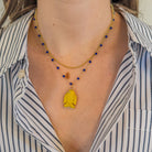 Person wearing a 14k gold necklace with lapis beads with a yellow agate fish pendant on the chain. It is paired with a 14k gold chain and wearing a striped shirt. 