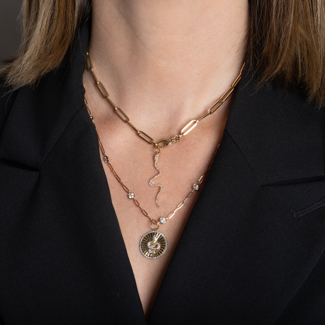 A person wearing a 14k gold paperclip chain necklace with a 14k gold diamond serpent charm layered with a 14k gold diamond station necklace and a 14k gold serpent charm.