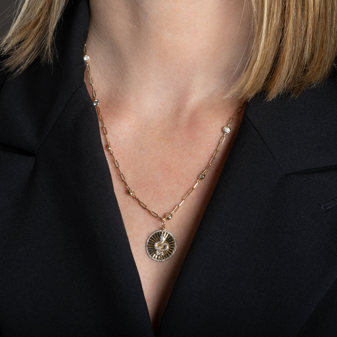 A person wearing a 14k gold diamond station paperclip necklace with a gold and diamond serpent pendant attached. 