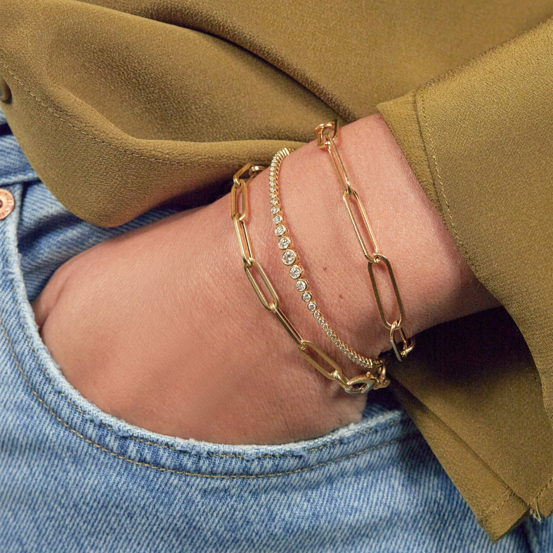 A person wearing two gold paperclip chain bracelets and a diamond flexi tennis bracelet.