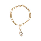 A gold paperclip chain bracelet with a diamond hamsa charm attached with a gold pendant bail laid out on a white background. 